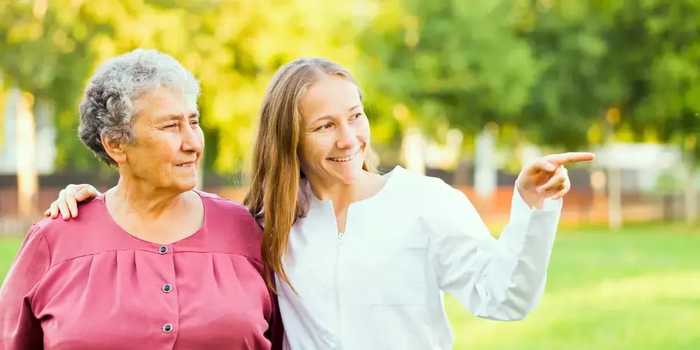 Smiling elderly woman and younger female caregiver enjoying a walk in the park, illustrating the importance of caring for elderly parents.