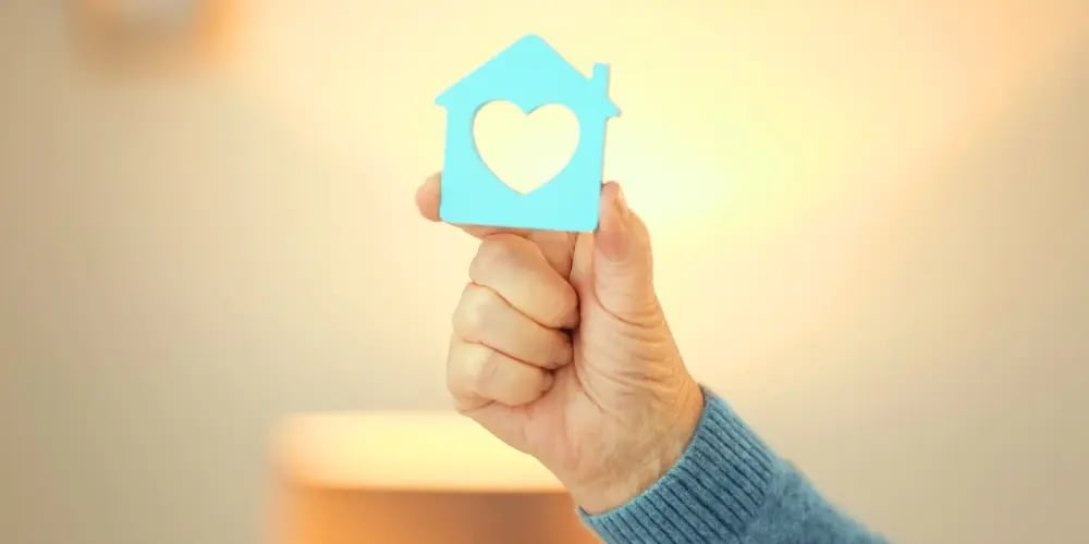 Hand holding a blue house icon with a heart cut-out, symbolising duty of care in aged care and a safe home environment.