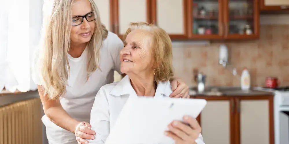 Caregiver assisting an elderly woman with a tablet in the kitchen, illustrating the process of arranging respite care for dementia patients.