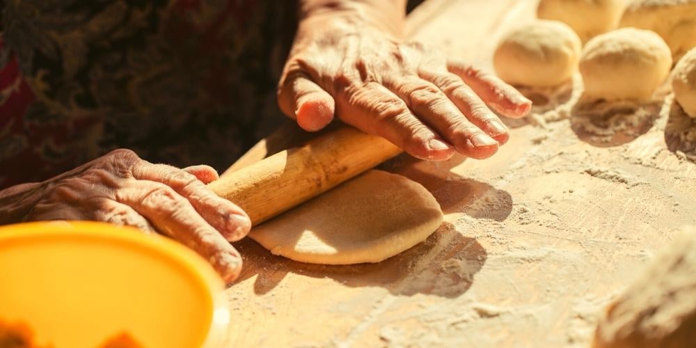 Photo of a senior person cooking, specifically rolling bread dough with a rolling pin.