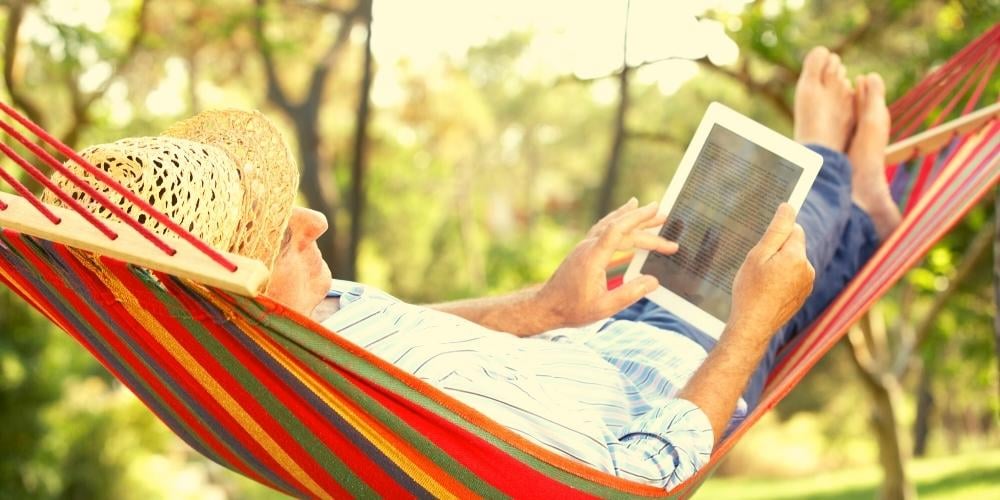 Senior citizen relaxing in a hammock in his garden reading computer game instructions on his iPad.