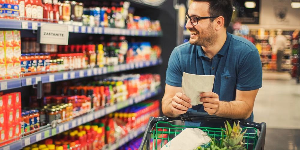 Smiling man with glasses holding a shopping list, navigating a supermarket aisle with a cart, exemplifying the assistance with grocery shopping provided by Level 2 Home Care Packages.