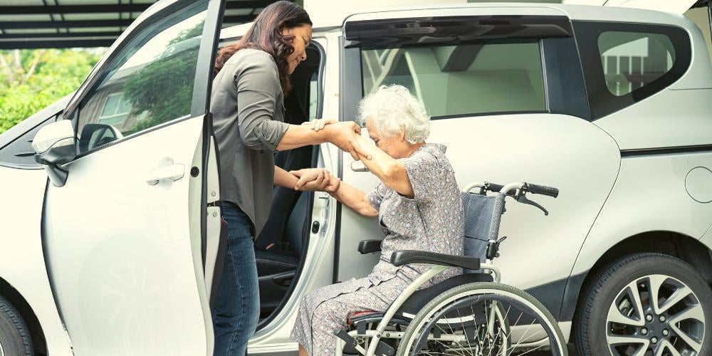Compassionate caregiver assisting an elderly woman in a wheelchair into a car, showcasing the transportation support for medical appointments included in Level 2 Home Care Packages.