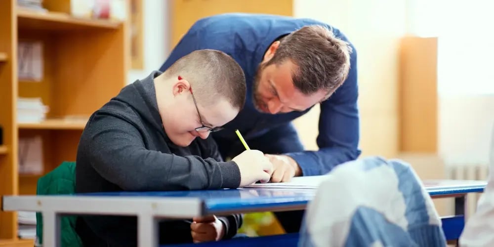 Boy with Down syndrome receiving guidance from a teacher, showing NDIS improved daily living and assistance with daily tasks.