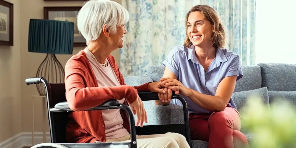 Personal care assistant talking with an elderly woman in a wheelchair, offering companionship and support in a comfortable home setting.