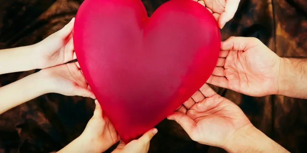Hands of multiple individuals holding a large red heart symbolising teamwork, compassion, and shared commitment in personal care services.