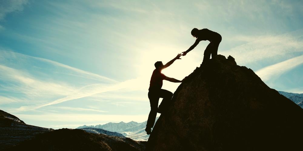 A couple climbing a mountain together offering a helping hand as they reach the summit.