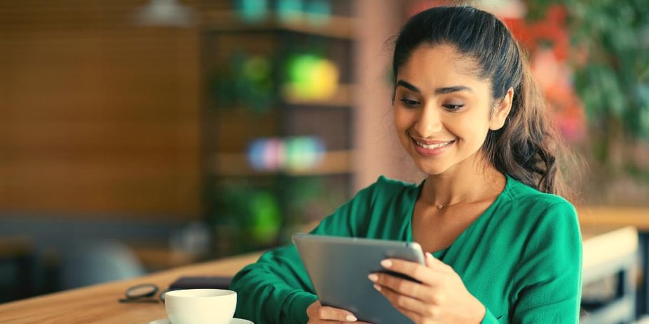 A lady in a cafe stood up with her coffee on the table beside her. She's browsing support coordinator jobs on her iPad.