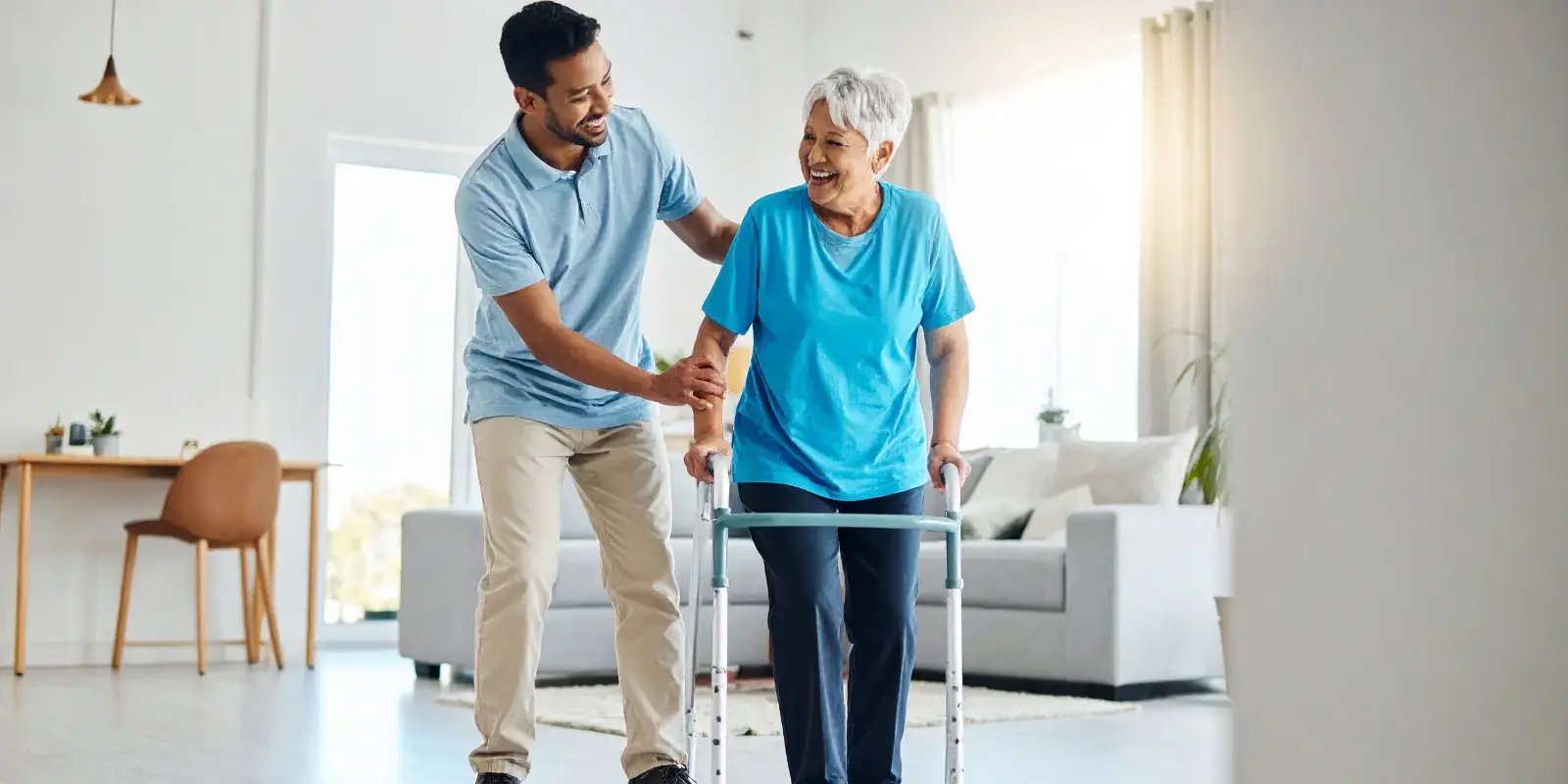 Male physiotherapist assists a smiling senior woman with a walker in her home, demonstrating an allied health service available via a Home Care Package.