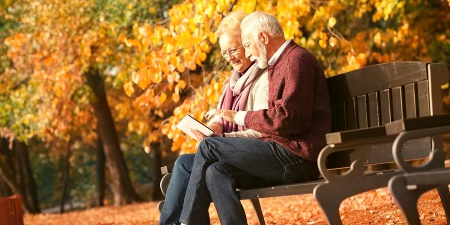 An elderly couple sitting on a park bench surrounded by trees with golden leaves. They have an iPad and are researching their aged care options together.