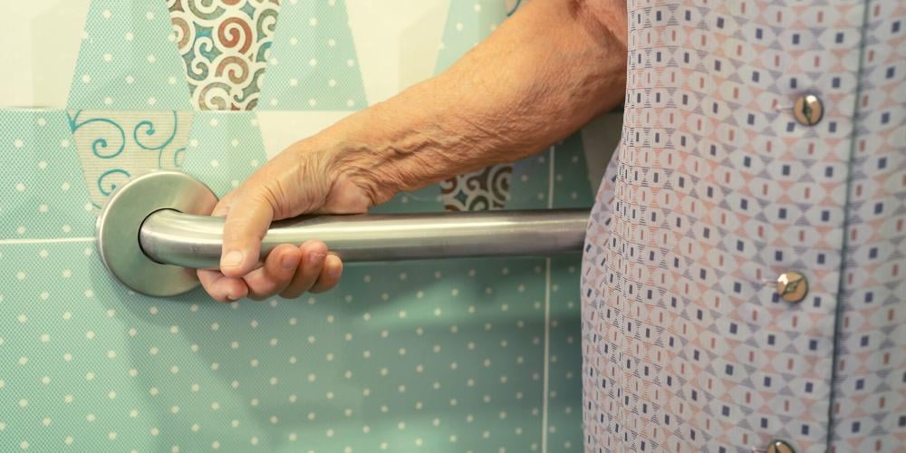 An elderly lady holding onto a hand rail in her bathroom for support. She has had the handrail fitted as an NDIS home modification.