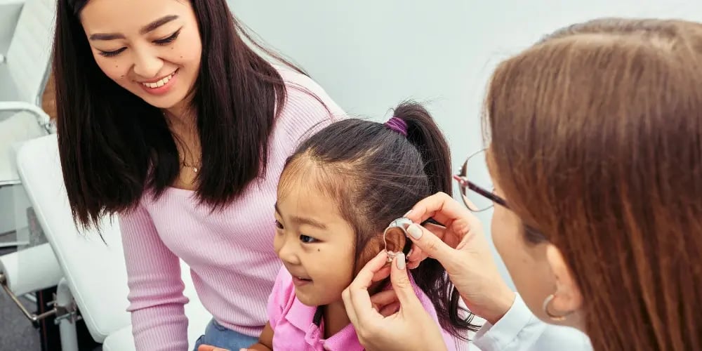 Child smiling as a clinician fits a hearing aid, with a parent observing, supporting the management of sensory hearing loss.