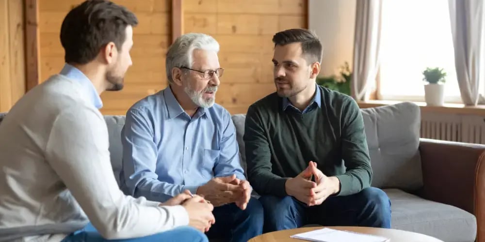 A family has a serious discussion about transitioning to in-home care, with two adult sons talking supportively with their elderly father about his needs.