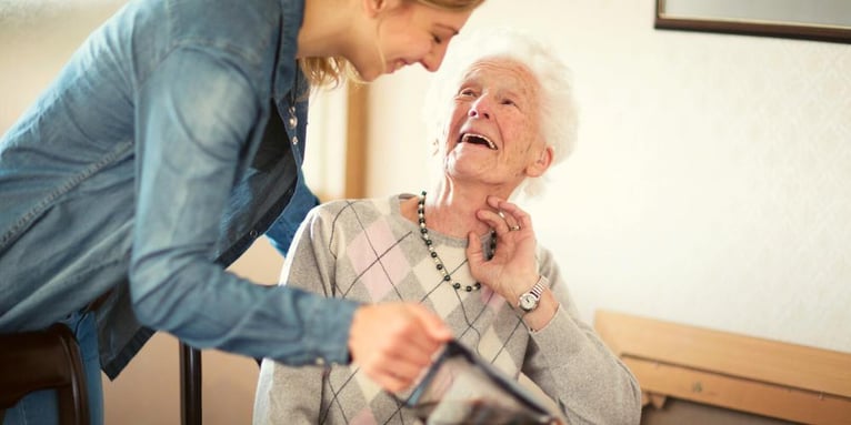 An elderly lady smiling and looking up at her professional carer who is pouring her a cup of tea.