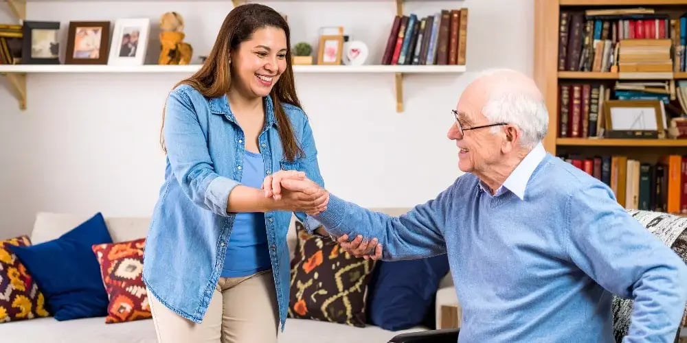 A friendly in-home caregiver providing mobility assistance to an elderly man, helping him stand up safely from his chair.