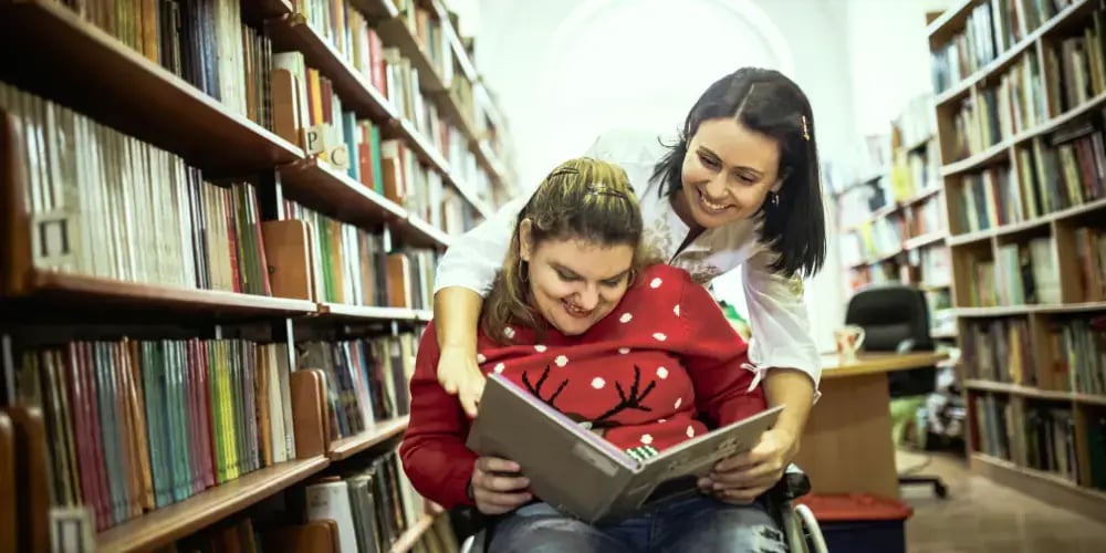 Caregiver helping a young woman in a wheelchair enjoy a book in a library, symbolising the assistance available through NDIS respite care services.