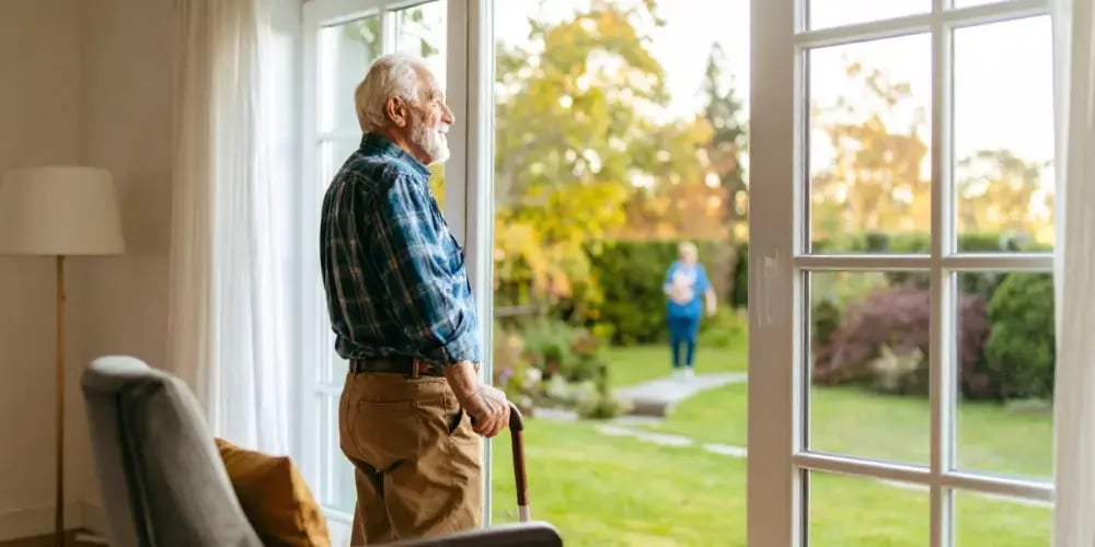 An elderly man with a walking cane stands comfortably in his home, looking out at his garden, thanks to a sustainable in-home care plan that supports his independence.
