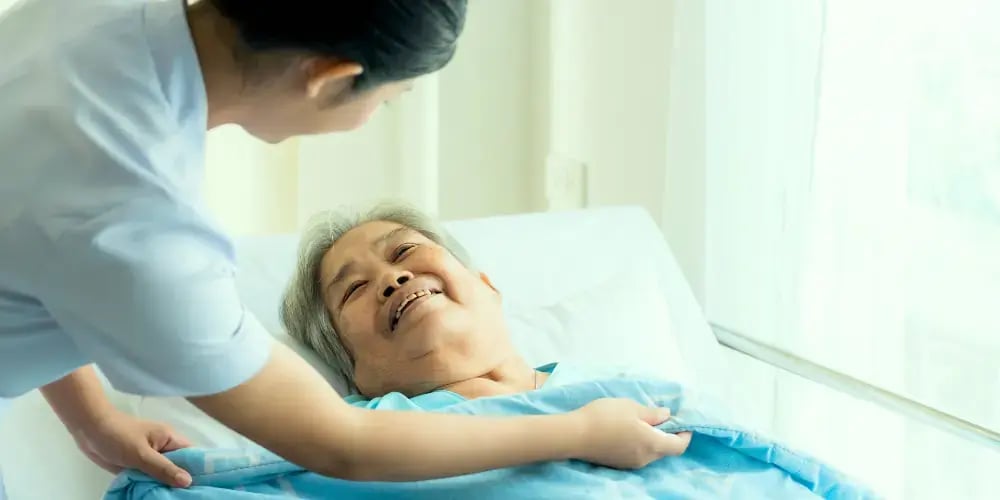 A smiling elderly man in bed being tenderly cared for by a nurse, illustrating personalised high care in aged care settings.