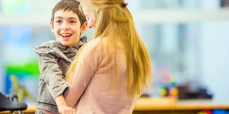 Smiling child with a disability being embraced by a supportive caregiver in a bright, indoor setting, demonstrating the comfort and support of respite care for people with a disability. This image highlights the importance of respite care for people with a disability, providing essential temporary relief to families and primary caregivers while maintaining a caring and safe environment for their loved ones.