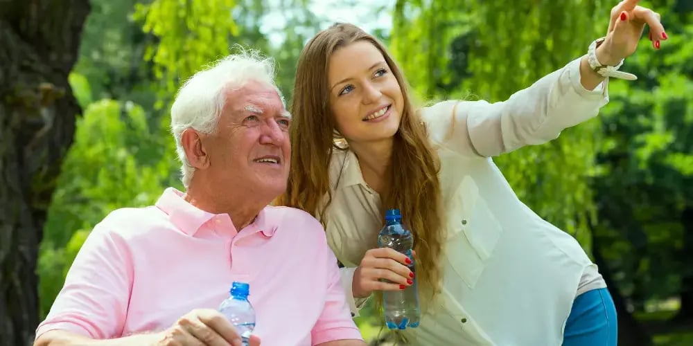 An elderly man with white hair, dressed in a pink polo shirt, is sitting outdoors, holding a bottle of water. Next to him is a younger woman with long brown hair, dressed in a white blouse and jeans, also holding a water bottle. She is pointing into the distance, and both are looking in that direction with interest. They are surrounded by lush green trees and nature. This image conveys the importance of respite care in providing enriching experiences and outdoor activities for the elderly, promoting their well-being and engagement.