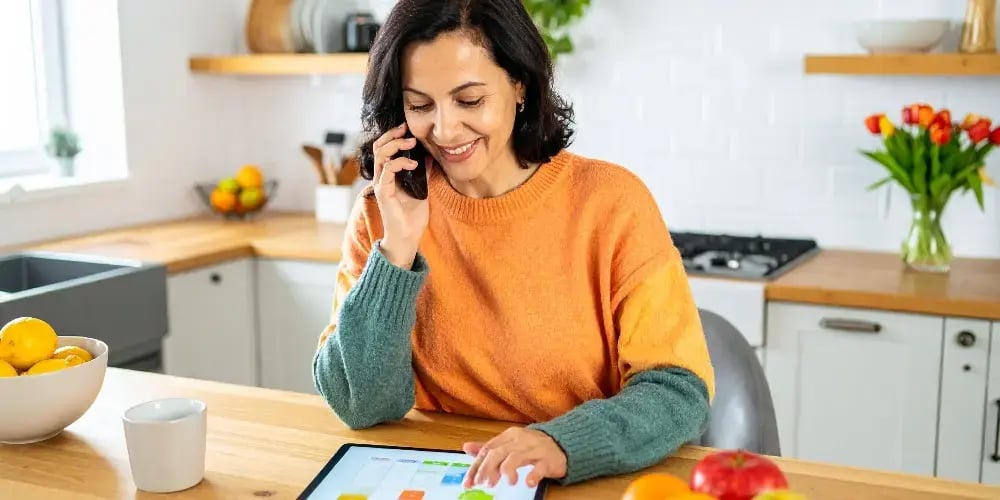 A woman proactively coordinates a sustainable in-home care plan, scheduling services on a tablet while talking on the phone with a care provider.